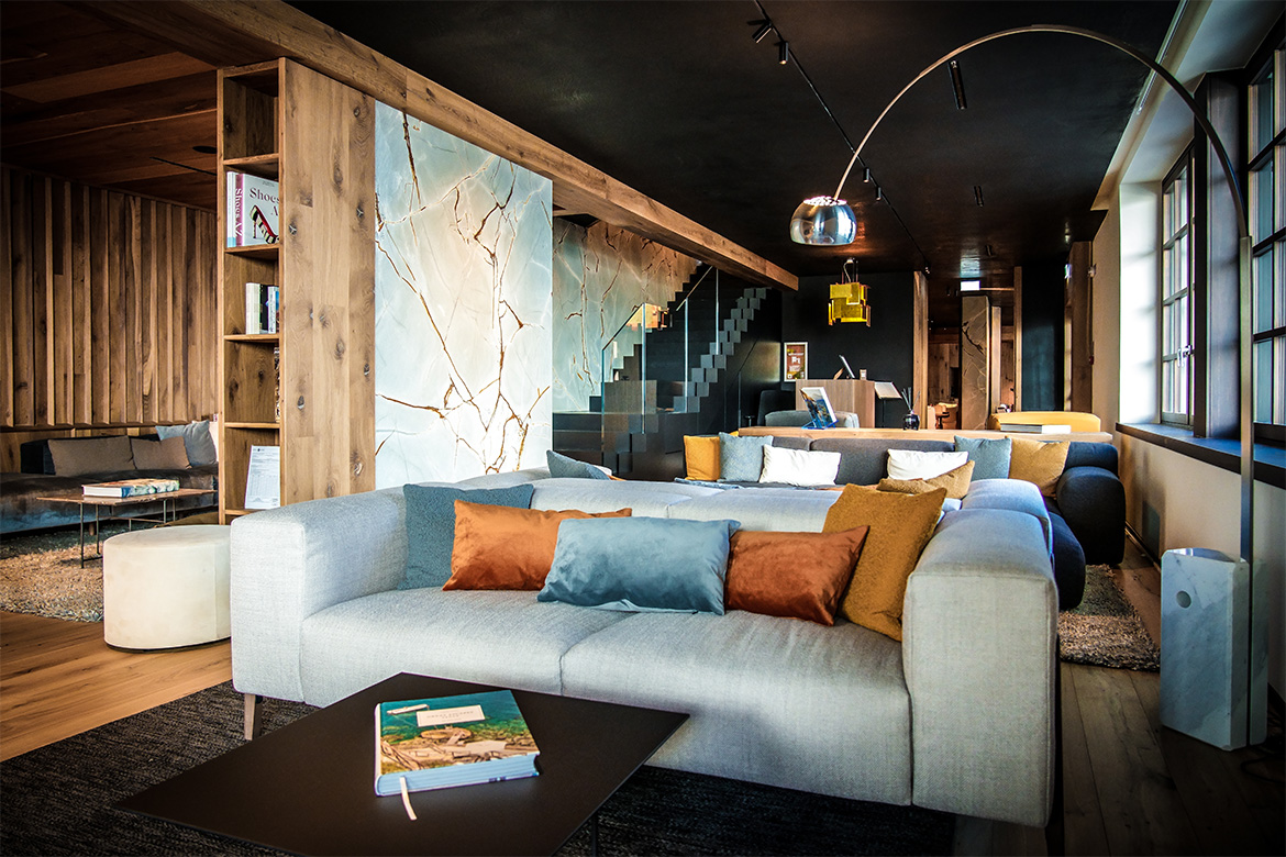 Lounge accogliente in Hotel Al Piolet con divani imbottiti, cuscini colorati, pareti in legno, dettagli effetto marmo e atmosfera calda da montagna.