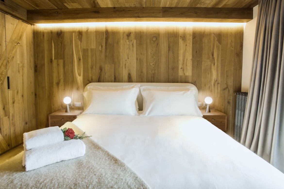 Camera matrimoniale accogliente in Hotel Al Piolet con pareti e testiera in legno, letto bianco, comodini coordinati, luci da lettura e arredi Pianca Contract.