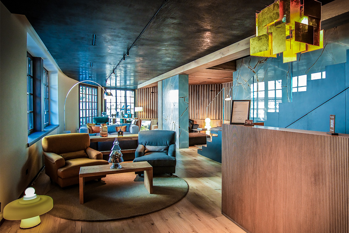Reception accogliente in Hotel Al Piolet con bancone in legno, area lounge, poltrone imbottite, illuminazione scenografica e atmosfera calda da montagna.