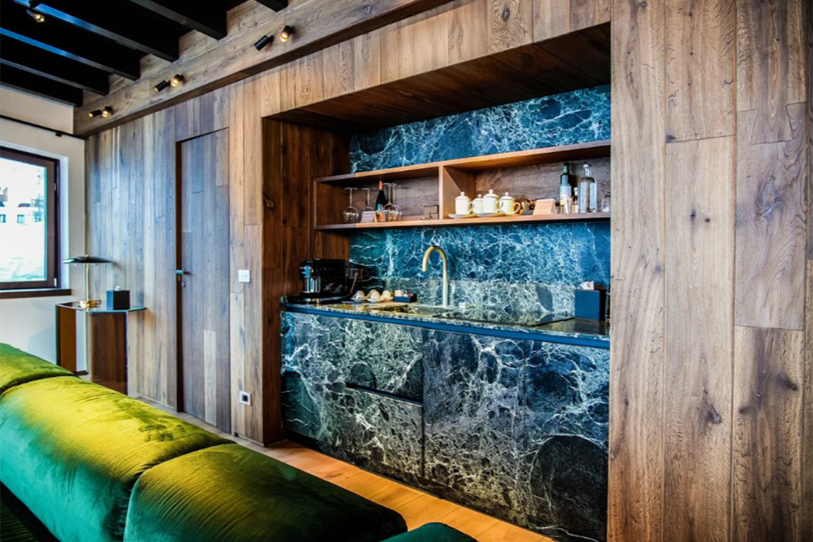 Camera moderna in Hotel Al Piolet con angolo cucina effetto marmo scuro, arredi in legno, divano verde e atmosfera calda da montagna.