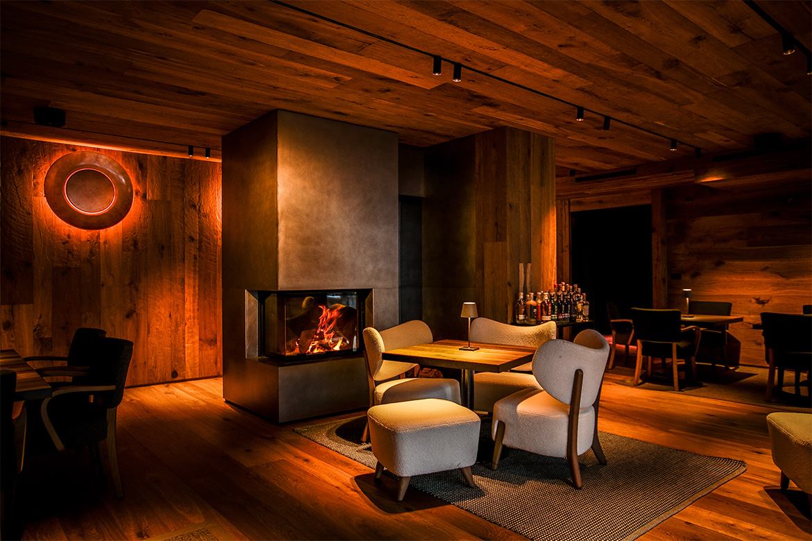 Lounge accogliente in Hotel Al Piolet con camino acceso, pareti e soffitto in legno, poltrone imbottite, tavolini e atmosfera calda da montagna.