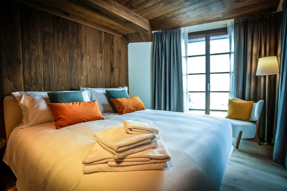 Camera matrimoniale accogliente in Hotel Al Piolet con pareti e soffitto in legno, letto bianco, cuscini colorati, poltrona di design e atmosfera calda da montagna.