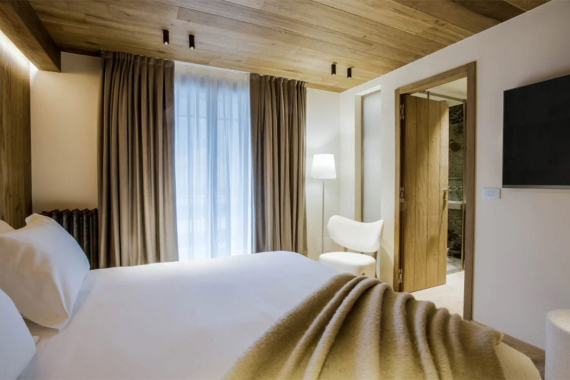Camera matrimoniale accogliente in Hotel Al Piolet con arredi in legno, letto bianco, tende chiare, poltrona di design, TV a parete e atmosfera calda da montagna.