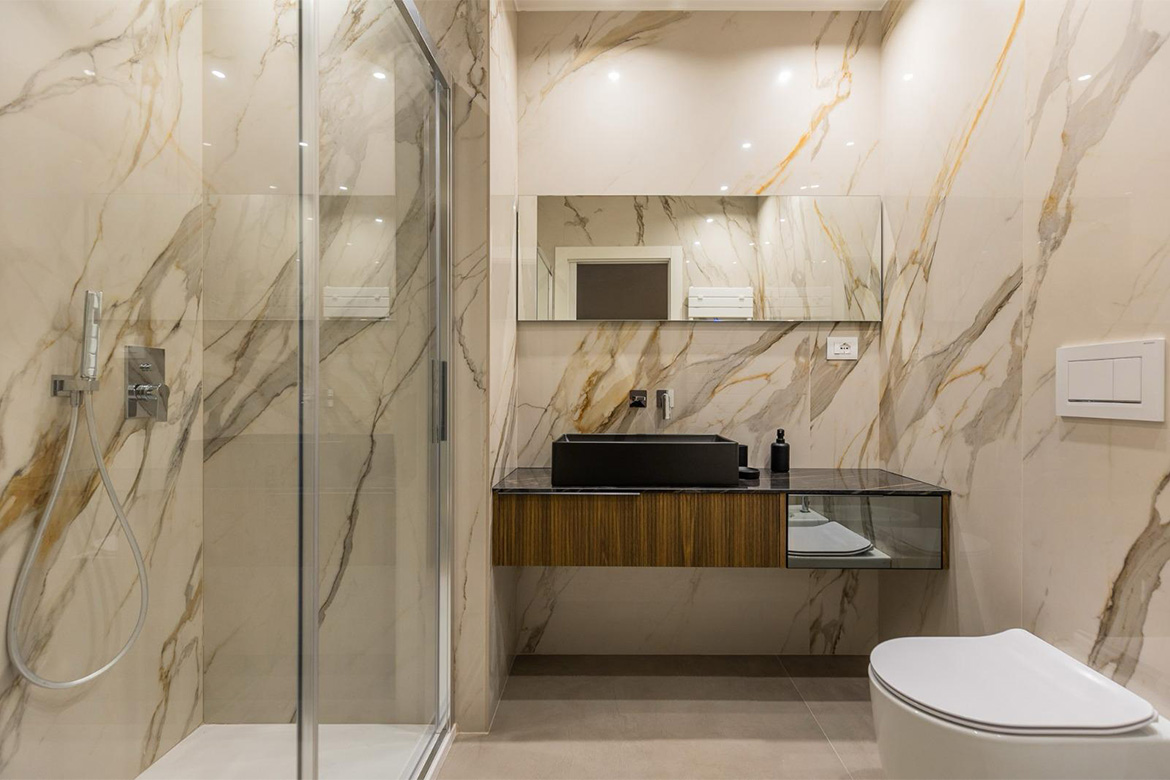 Bagno moderno con doccia in vetro, mobile sospeso in legno, lavabo nero, sanitari bianchi e rivestimento effetto marmo per B&B San Martino.