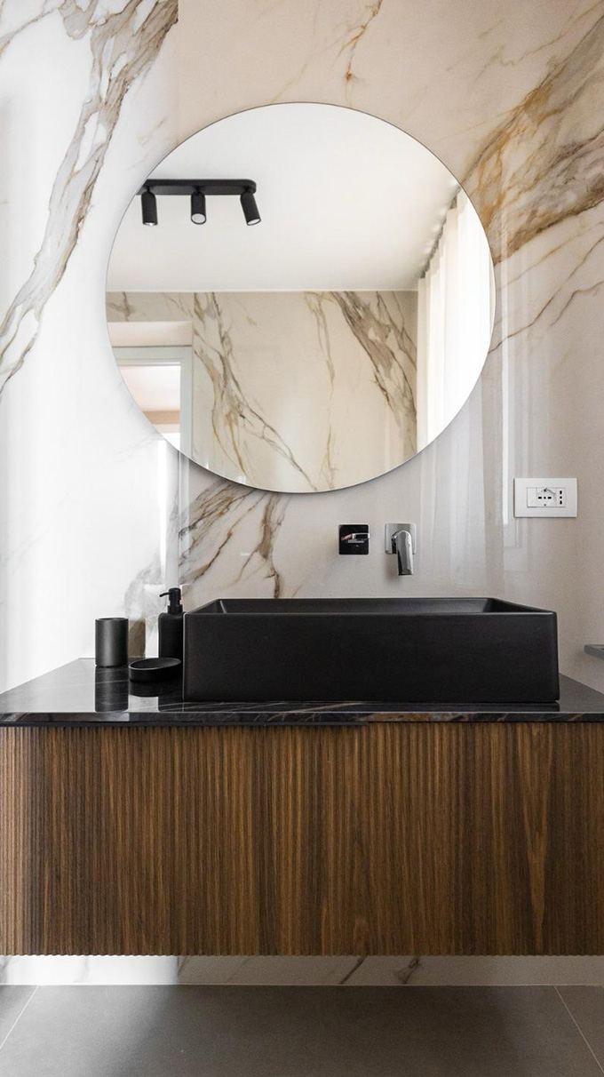 Bagno moderno con mobile sospeso in legno, lavabo nero, specchio rotondo e rivestimento effetto marmo, arredato da Pianca Contract per B&B San Martino.