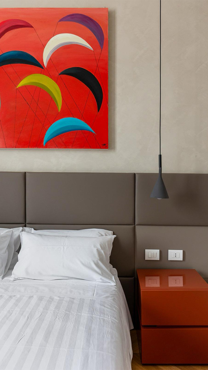 Camera da letto moderna con testiera imbottita, comodino rosso laccato, lampada a sospensione e quadro colorato sopra il letto.
