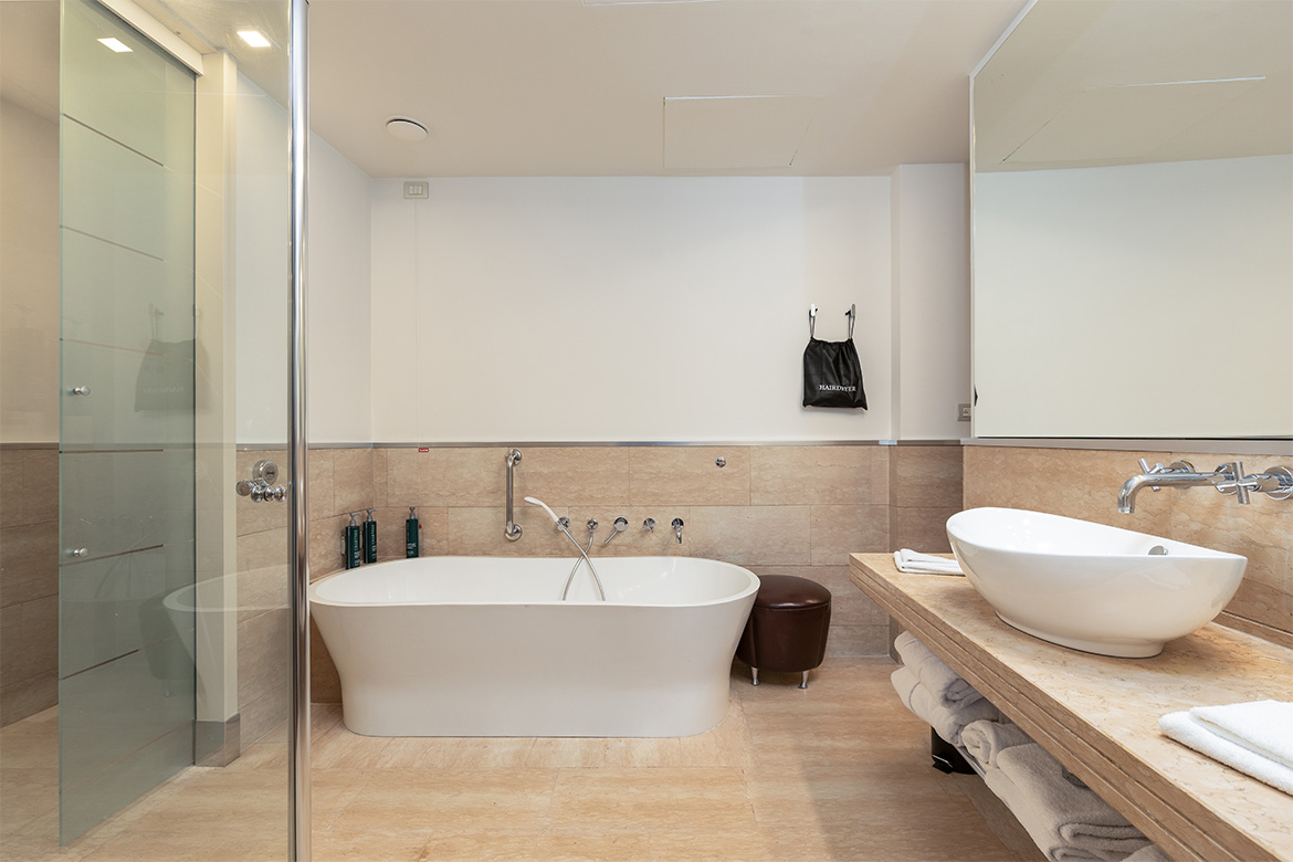 Bagno moderno in DoubleTree Hilton con vasca freestanding, doccia in vetro, lavabo da appoggio, piano effetto marmo e arredi Pianca Contract.