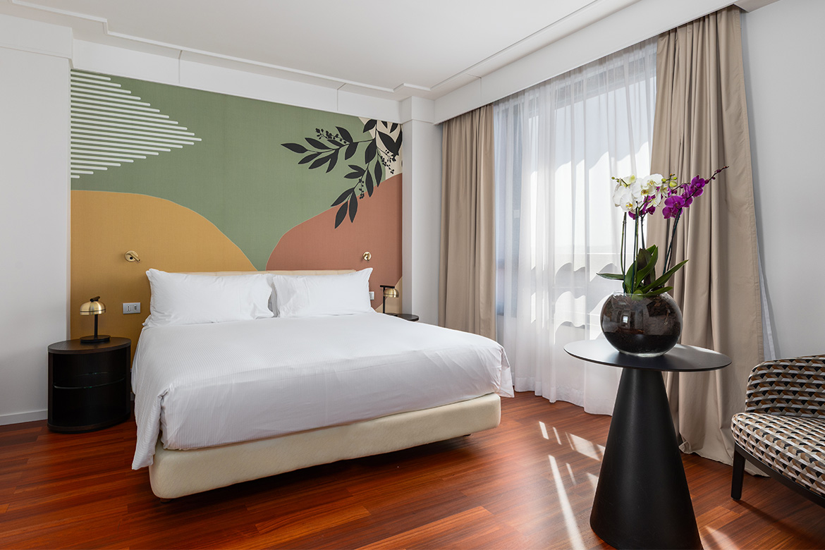 Camera matrimoniale moderna in DoubleTree Hilton a Firenze con letto imbottito, parete decorativa botanica, pavimento in legno, tavolino nero con orchidee e arredi Pianca Contract.