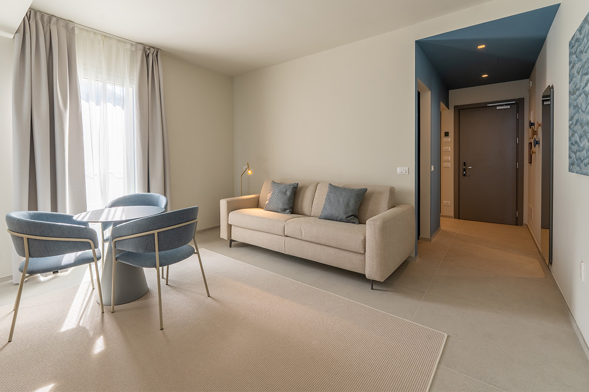 Zona living moderna in hotel vicino al mare con divano beige, tavolo rotondo, poltroncine azzurre, tende chiare e arredi Pianca Contract.