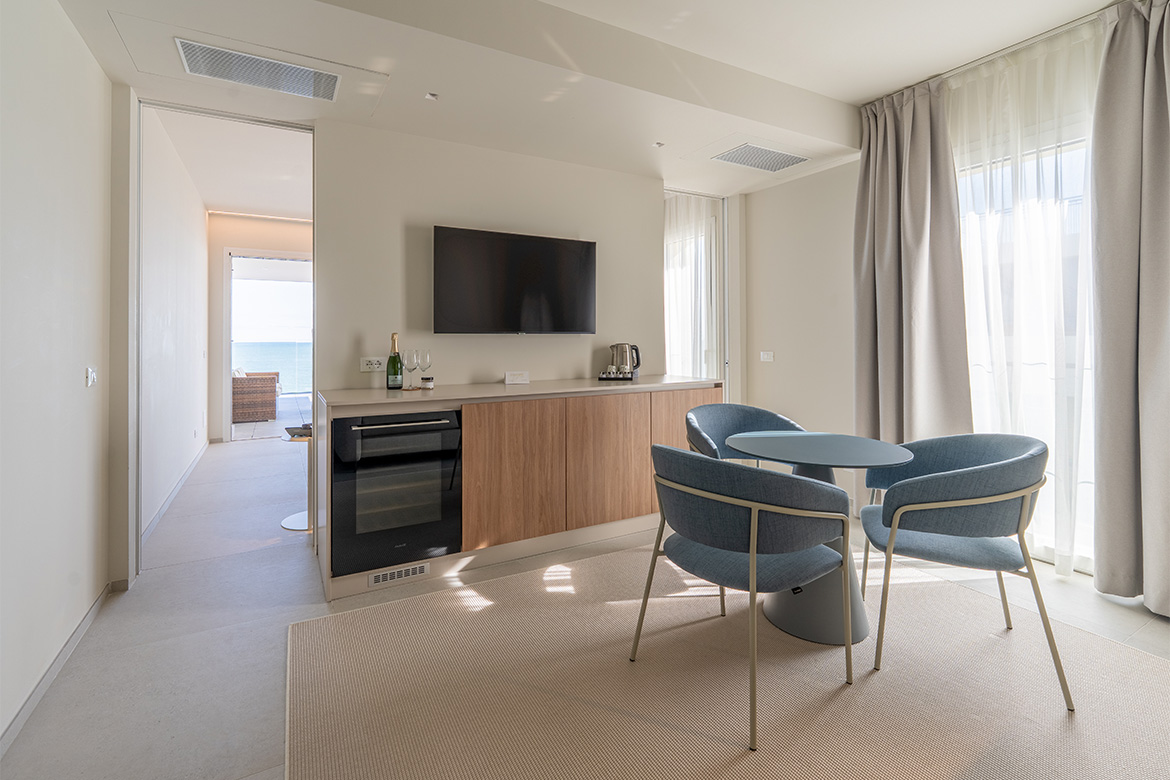 Zona living moderna in hotel vicino al mare con mobile TV, arredi in legno, tavolo rotondo, poltroncine azzurre, tende chiare e vista verso il balcone.