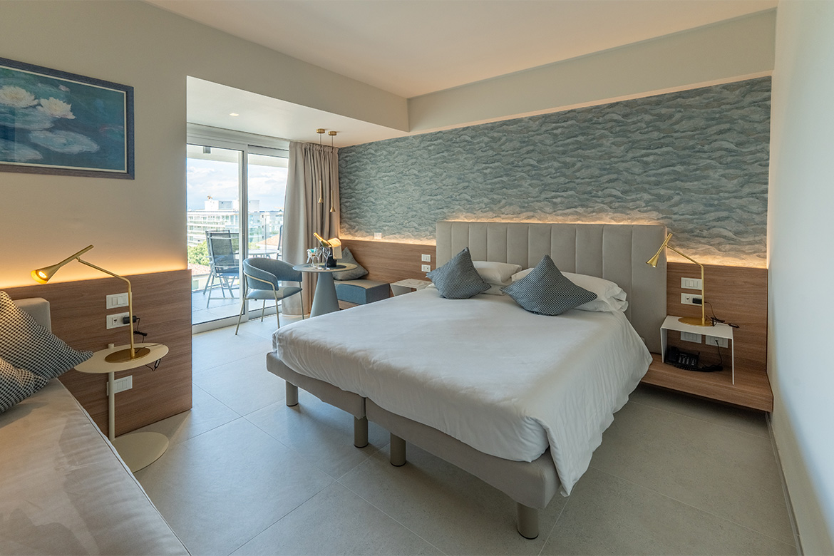 Camera moderna in hotel vicino al mare con letto matrimoniale imbottito, parete decorativa azzurra, arredi in legno, lampade da lettura e balcone, arredata da Pianca Contract.
