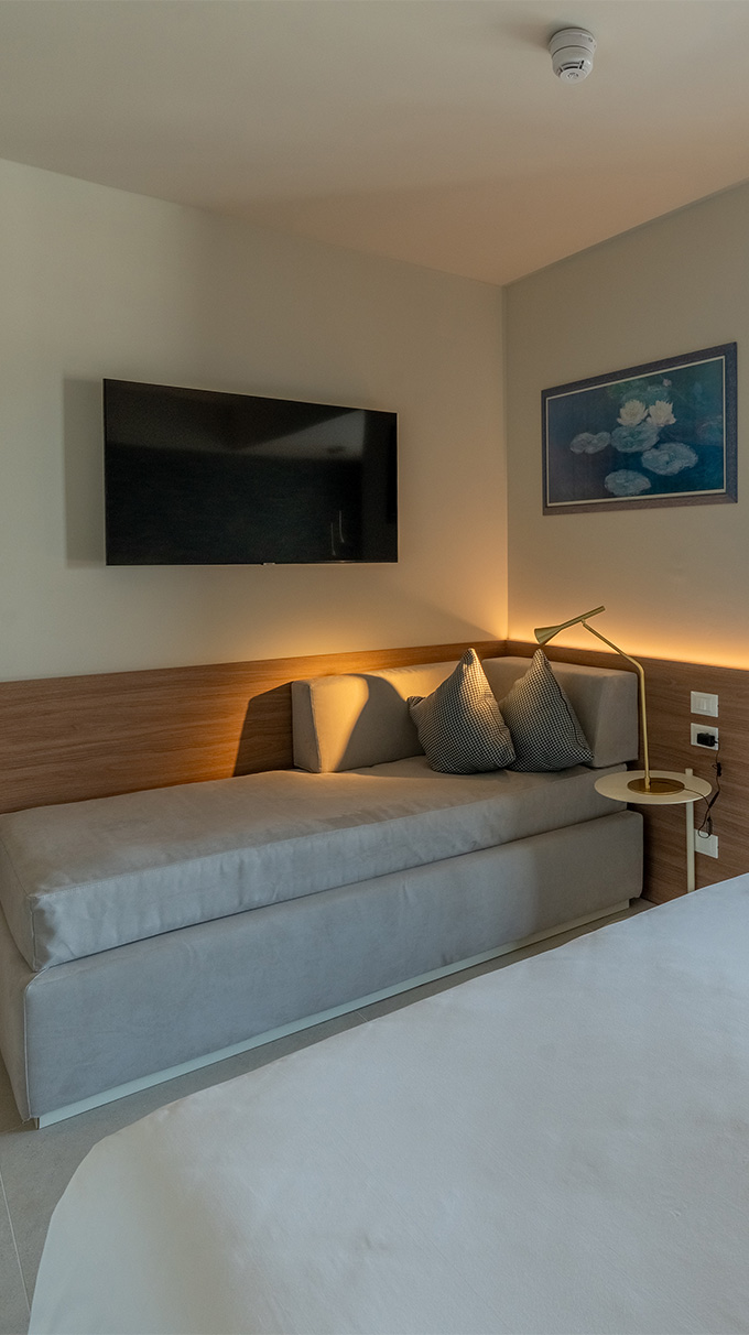 Camera moderna in Hotel Victoria con divano letto imbottito, mobile TV sospeso, illuminazione calda, comodino con lampada e arredi Pianca Contract.