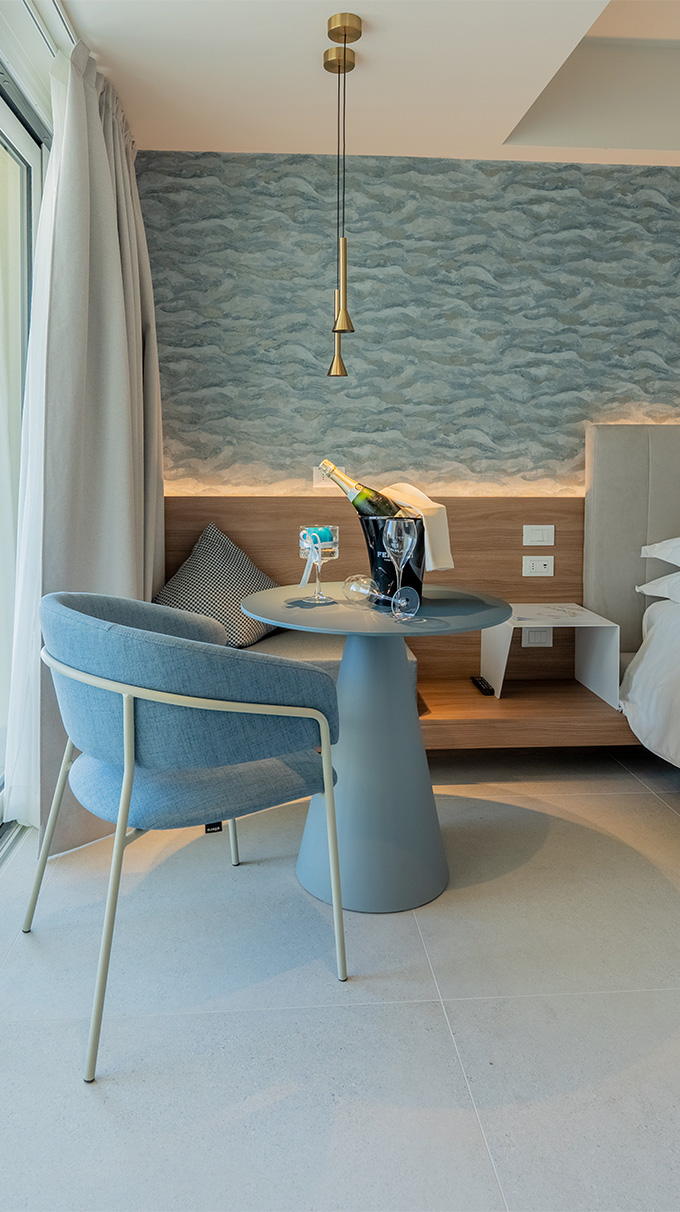 Angolo relax moderno in camera Hotel Victoria con poltroncina azzurra, tavolo rotondo, lampade sospese, parete decorativa azzurra e arredi Pianca Contract.