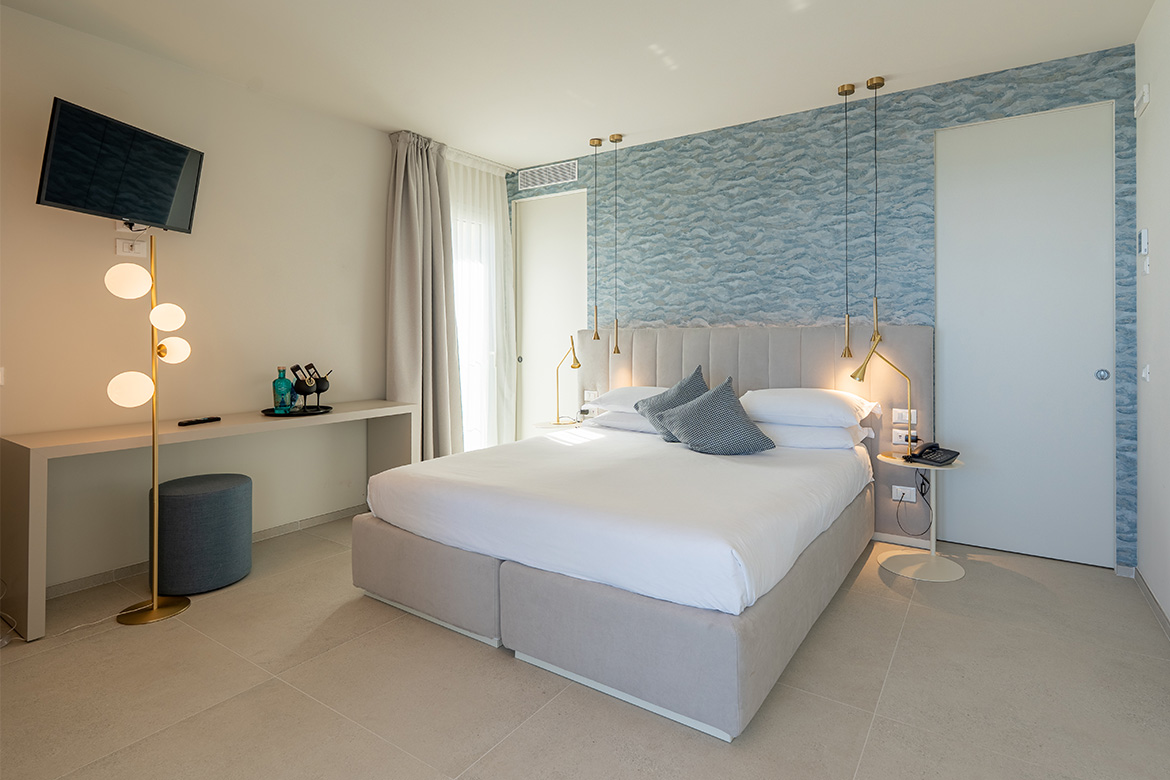 Camera moderna in hotel vicino al mare con letto matrimoniale imbottito, parete decorativa azzurra, lampade sospese, scrittoio e arredi Pianca Contract.