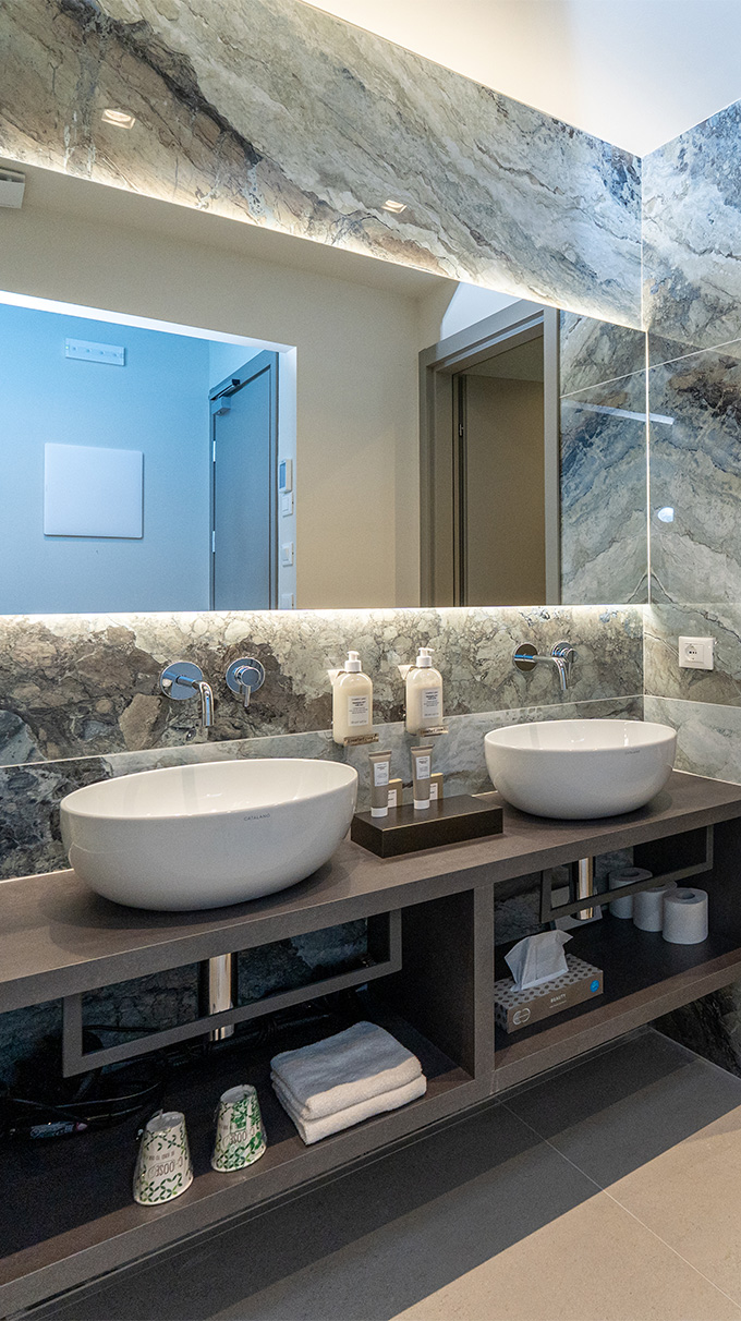 Bagno moderno in hotel vicino al mare con doppio lavabo da appoggio, grande specchio retroilluminato, mobile sospeso e rivestimento effetto pietra.