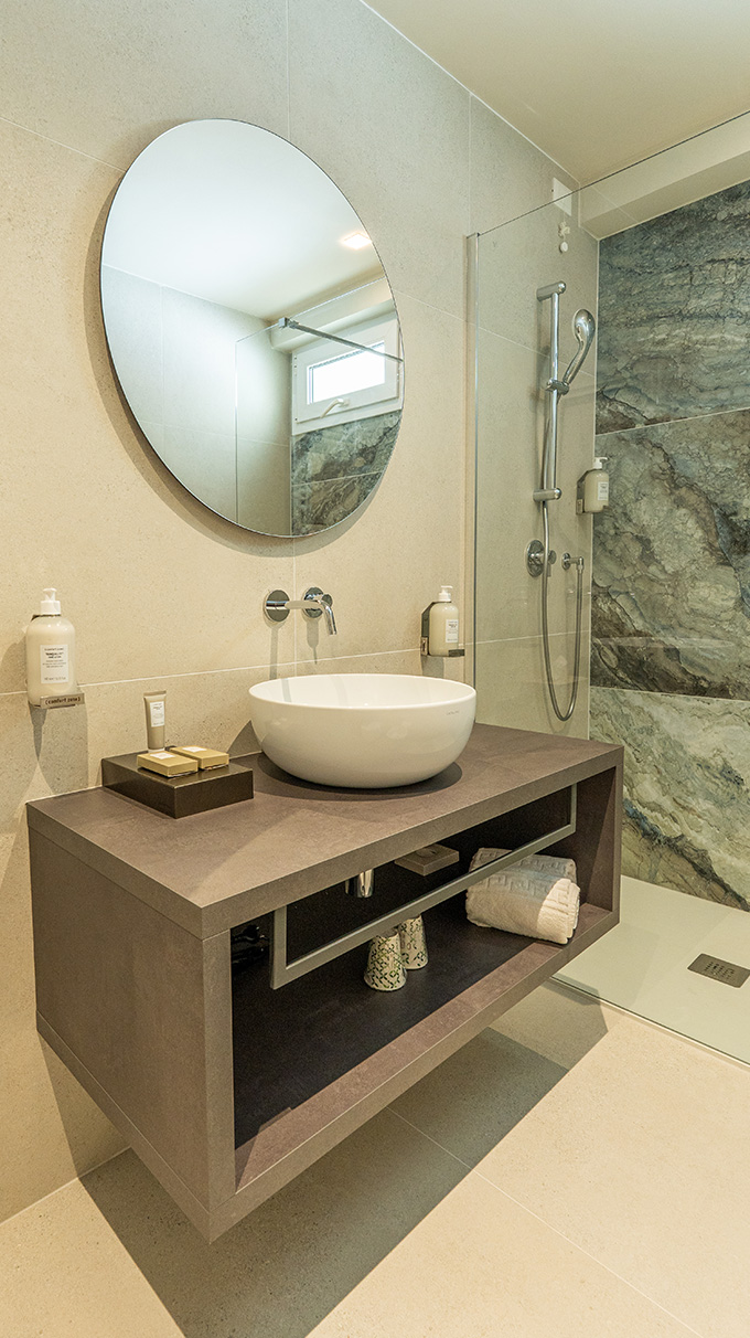 Bagno moderno in hotel vicino al mare con mobile lavabo sospeso, lavabo da appoggio bianco, specchio rotondo, doccia in vetro e rivestimento effetto pietra.