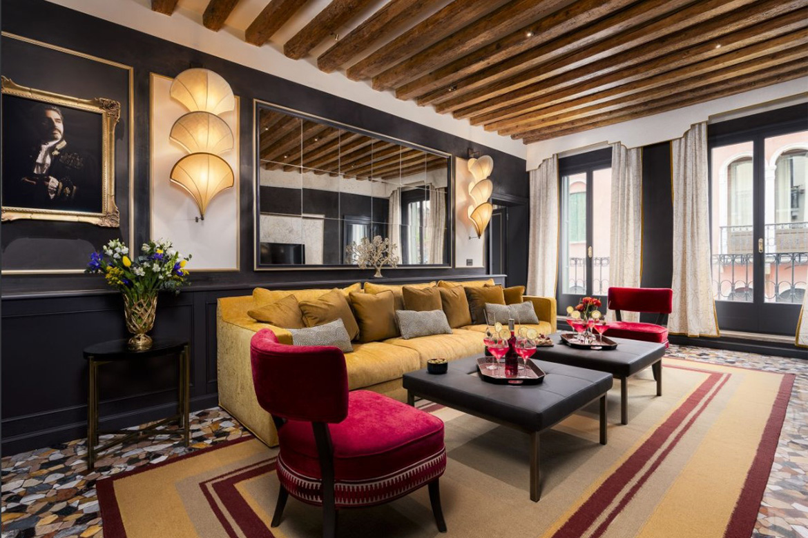Living elegante in hotel veneziano con divano giallo, poltrone rosse, tavolini centrali, travi in legno a vista e arredi Pianca Contract.