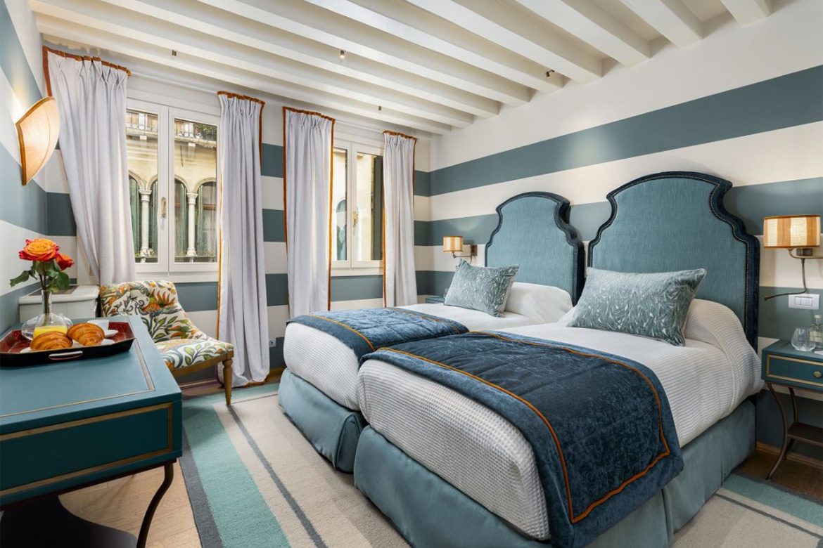 Camera doppia elegante con due letti singoli, testiere imbottite azzurre, pareti a righe, arredi coordinati e dettagli decorativi in stile hospitality.