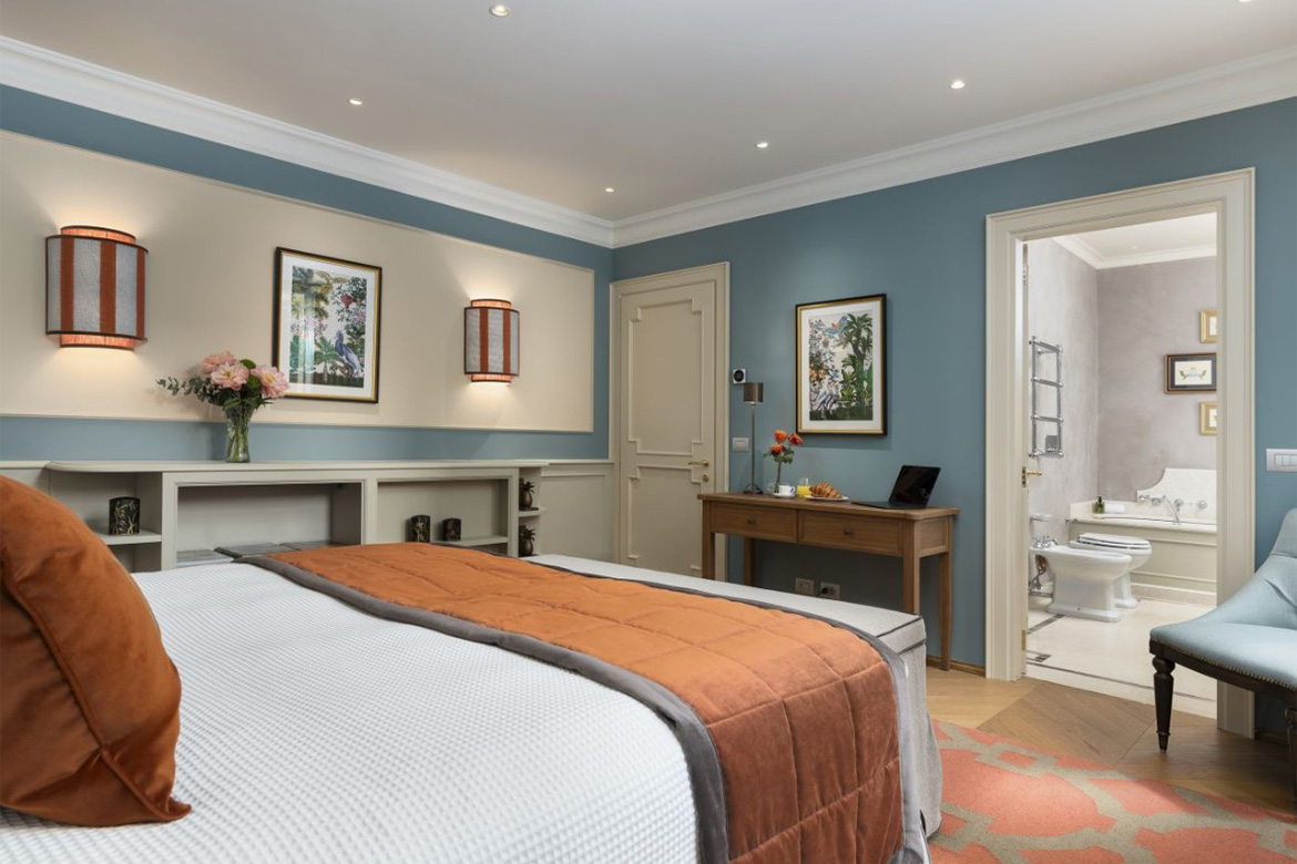 Camera matrimoniale elegante in hotel veneziano con letto imbottito, dettagli arancioni, pareti azzurre, scrittoio in legno e bagno en suite arredato da Pianca Contract.