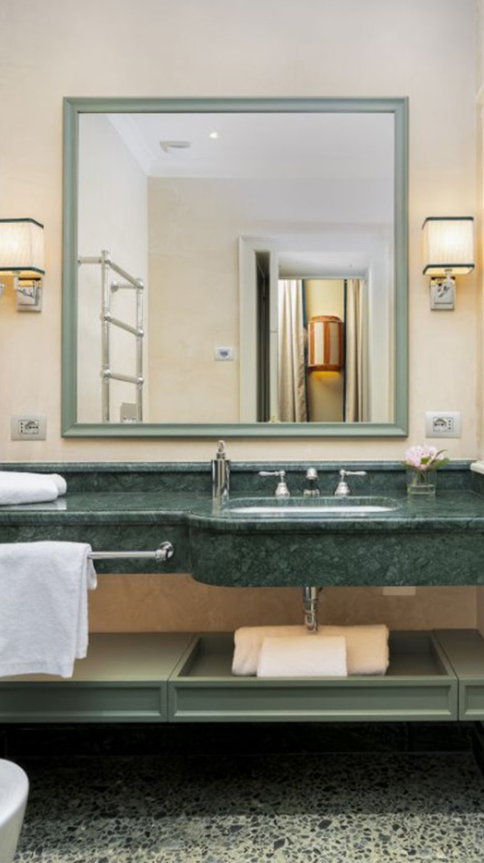 Bagno elegante in hotel veneziano con lavabo in marmo verde, grande specchio rettangolare, applique laterali e dettagli raffinati Pianca Contract.