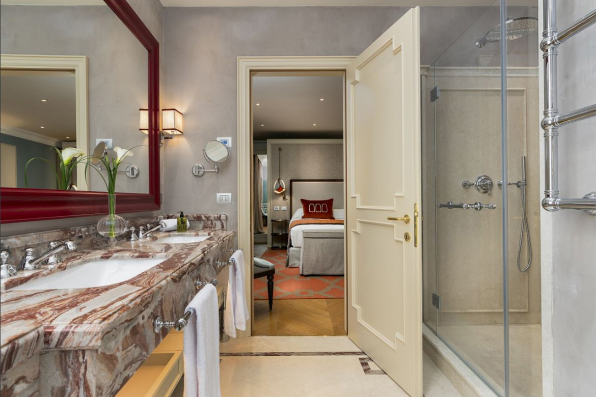 Bagno elegante in hotel veneziano con lavabo in marmo, grande specchio rosso, doccia in vetro e dettagli raffinati, arredato da Pianca Contract.