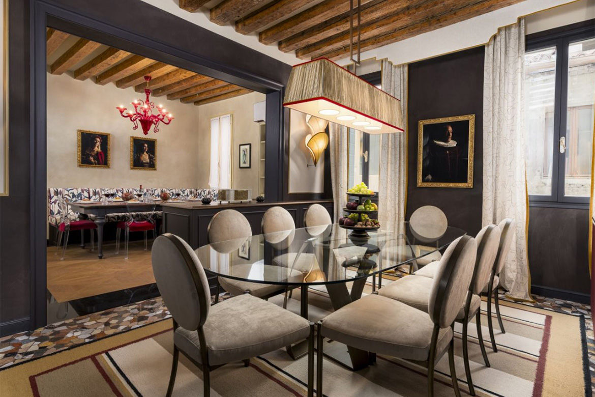 Sala da pranzo elegante in hotel veneziano con tavolo in vetro, sedie imbottite, lampada sospesa, travi in legno a vista e arredi Pianca Contract.