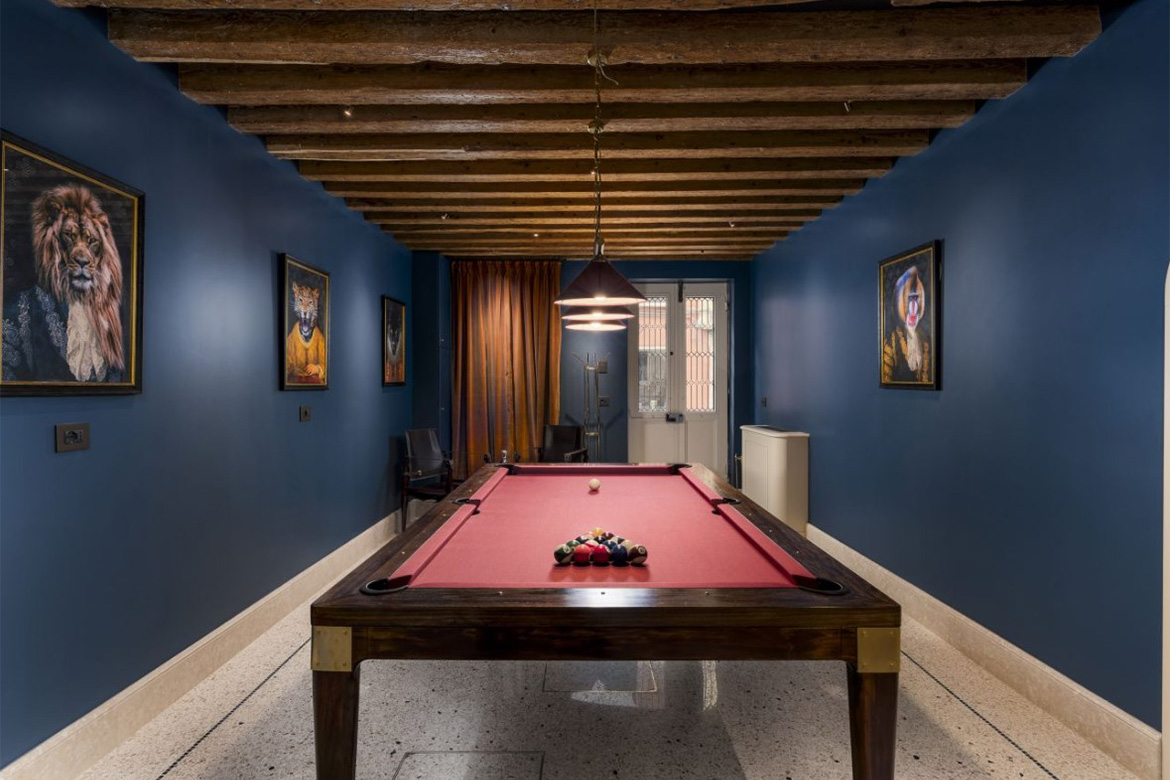 Sala biliardo elegante in hotel veneziano con tavolo da biliardo, pareti blu, travi in legno a vista, lampada sospesa e arredi Pianca Contract.