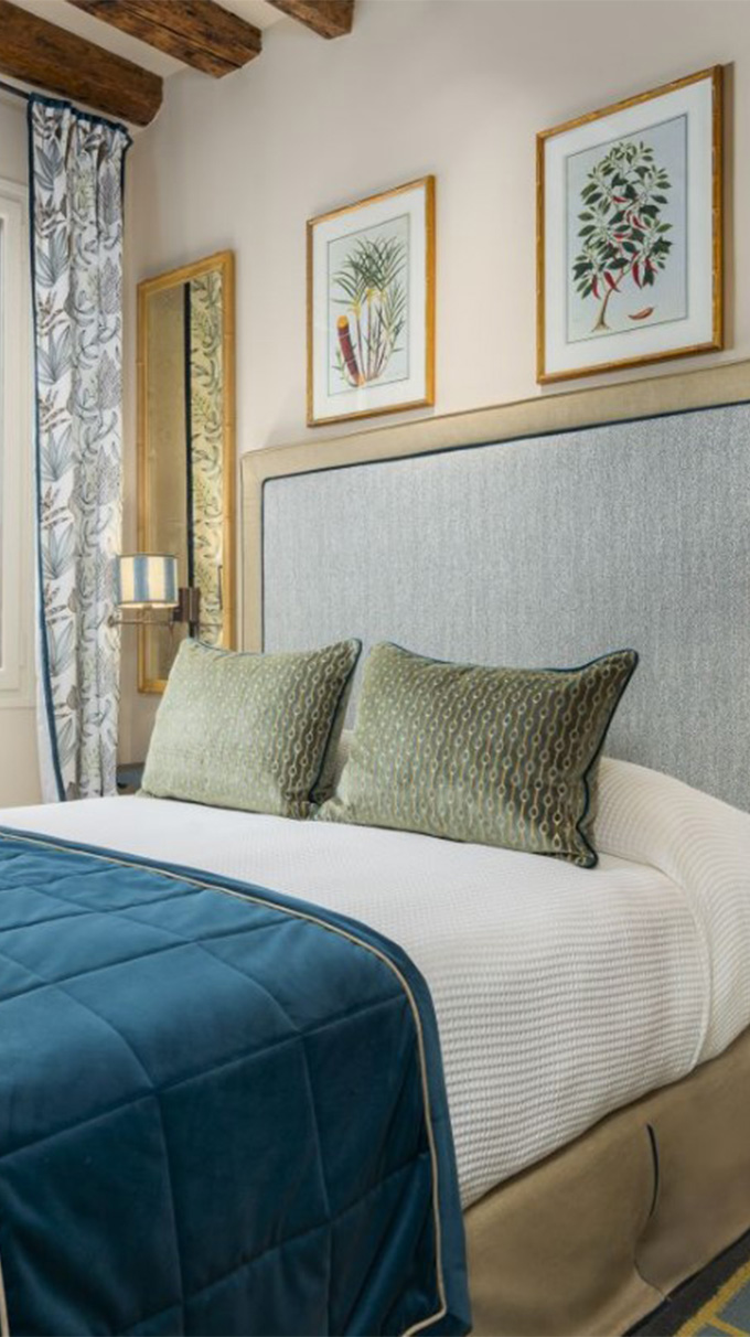 Camera matrimoniale elegante in hotel veneziano con letto imbottito, testiera grigia, cuscini decorativi, quadri botanici, travi in legno a vista e arredi Pianca Contract.