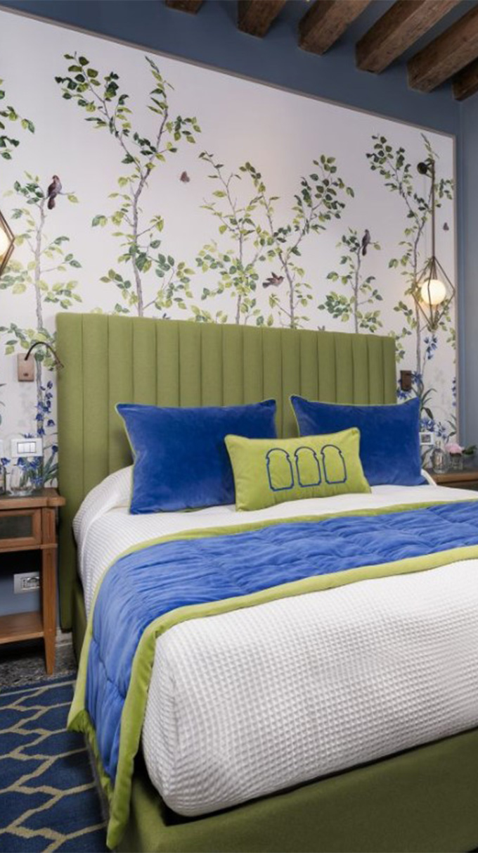 Camera matrimoniale elegante in hotel veneziano con letto imbottito verde, cuscini blu, carta da parati botanica, travi in legno a vista e arredi Pianca Contract.