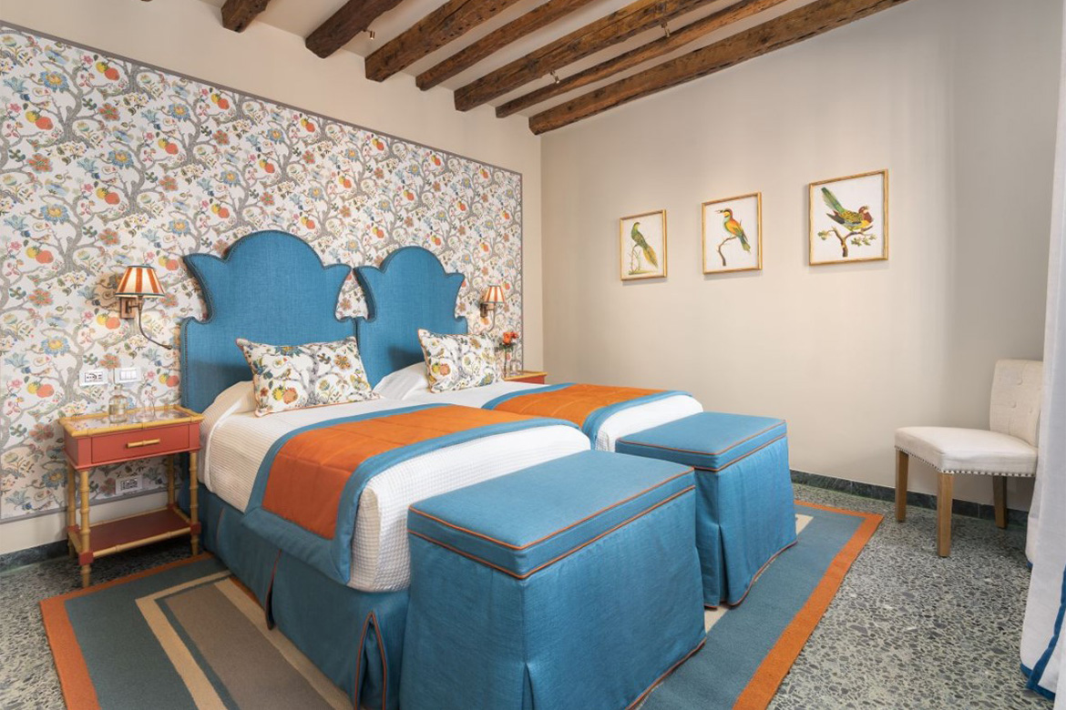 Camera doppia elegante in hotel veneziano con letti singoli, testiere imbottite azzurre, carta da parati floreale, travi in legno a vista e arredi Pianca Contract.